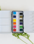 The Faire Travel Watercolor Paint Palette, featuring twelve vibrant pans, sits open on a white surface surrounded by art supplies, green leaves, and yellow-and-white flowers.