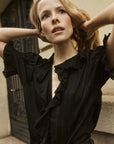 A woman with wavy blonde hair wears the DOEN Henri Top—an elegant black Edwardian blouse—as she stands outside, looking upward with her hands behind her head, set against a neutral-toned background with a decorative door.