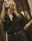 A woman with long wavy blonde hair wears the DOEN Henri Top, featuring lace-trimmed ruffles, and high-waisted dark jeans. She stands before an ornate metal gate and yellow textured wall, gazing upward with a relaxed expression.