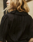 A person with wavy blonde hair wears the DOEN Henri Top, featuring an Edwardian-inspired ruffled collar, seen from behind and slightly in profile near a beige wall.