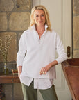 A person with blonde hair exudes a preppy vibe indoors, wearing a white collared Patrick Popover Henley by Frank & Eileen over a 100% cotton shirt and olive pants. They're leaning against a wooden chair, set against a stone wall with a table holding books and a vase in the background.