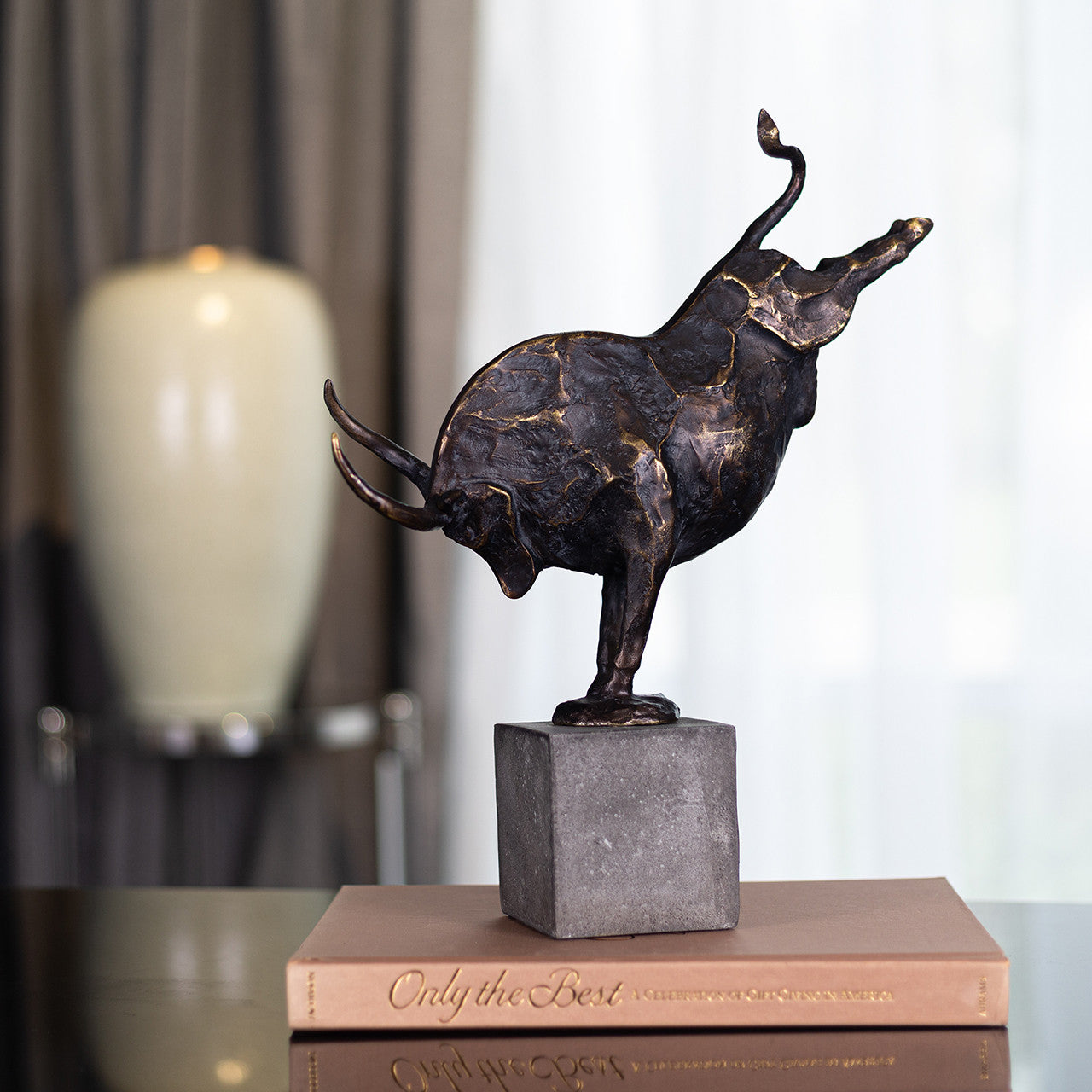 The Import Collection’s Bull Statuary, ideal for home decor, stands on one front leg atop a gray stone base and a closed book titled “Only the Best.” A blurred lamp and curtain add elegance to the background.