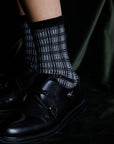 A close-up of a person’s lower leg wearing Maria La Rosa Iceland Socks—women’s mid-calf, black and white plaid merino wool—paired with black leather double monk strap dress shoes, set against a dark textured background.