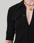 A person wears the Citizens Of Humanity/AGOLDE Amina Polo Dress—a black, ribbed cotton button-up with half sleeves and a chest pocket—paired with layered gold chain and coin pendant necklaces. The background is plain light gray.