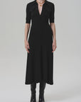 A woman in the Amina Polo Dress by Citizens Of Humanity/AGOLDE—fitted, black, with chest pockets—pairs it with a gold necklace and knee-high black boots against a plain light gray background.