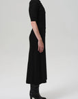 A person stands in profile against a plain background, wearing the Citizens Of Humanity/AGOLDE Amina Polo Dress—an ankle-length black button-down with short sleeves and a collar—paired with black heeled boots and arms resting at their sides.