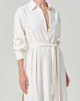 A woman wears the Camillia Shirt Dress in Portofino Twill by Citizens Of Humanity/AGOLDE—a long-sleeved, collared white dress with a button-down front, tie waist, and chest pocket. She accessorizes with a gold necklace; her face is partially out of frame.