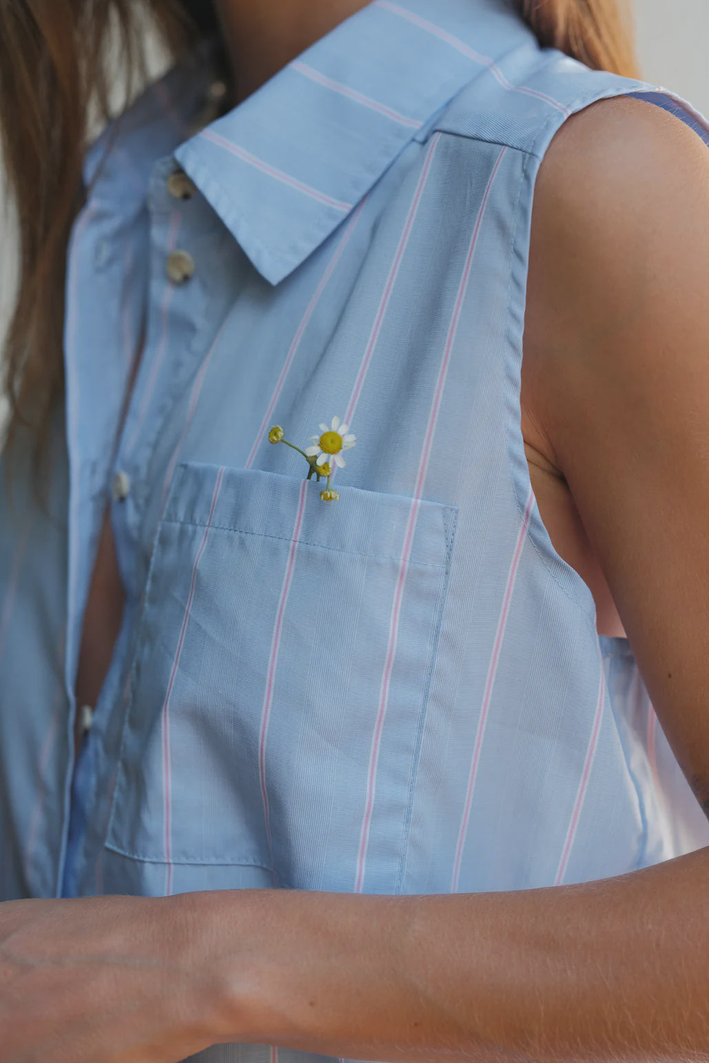 Close-up of a woman in a light blue cotton Donni Pop sleeveless shirt, with three small daisies tucked in her shirt pocket, focusing on the textured fabric and the simple elegance of the flowers.