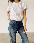 A person wearing "The Crew" plain white 100% cotton T-shirt by Hey Gang and blue jeans stands against a neutral backdrop, one hand casually in the pocket. The relaxed pose highlights the enzyme-washed style of this vintage find.