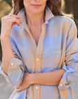 A woman stands outdoors in the Frank & Eileen Eileen Button Up Shirt F25—a relaxed fit with bust-flattering buttons and a light blue, beige, and gray plaid—arms crossed, smiling slightly against a softly blurred backdrop.