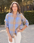 A woman with wavy brown hair stands outdoors in the Frank & Eileen Eileen Button Up Shirt F25, featuring bust-flattering button placement and a relaxed fit, paired with white pants amid green bushes and trees.