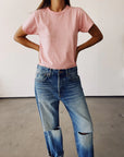 A person confidently poses with hands on hips wearing Hey Gang's 100% cotton vintage pink "The Crew" tee and ripped blue jeans, set against a plain, light-colored wall that highlights the casual yet stylish appeal of this enzyme-washed look.