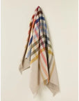 The Faire BM Merino Lamb Throw/Blanket, in beige with colorful red, blue, yellow, and green plaid stripes and fringed edges, is displayed neatly hanging on a light-colored wall.