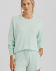 A woman wears the One Grey Day Warwick Pullover in light mint green with dorito stitching and matching shorts, standing with one hand in her pocket against a plain white background.