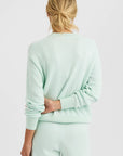 A woman with blonde hair in a loose bun is shown from behind, wearing the light mint green Warwick Pullover by One Grey Day and matching shorts. One arm is bent behind her back against a plain white background.