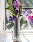 A Vintage Tall Tapered Pitcher by Faire, filled with purple and pink flowers, sits on a windowsill as sunlight streams in through the window behind it.