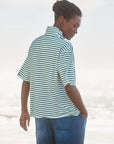 A woman stands outdoors with her back turned, wearing the Frank & Eileen Patty Popover SS Henley in mint-green and white stripes with blue pants. One hand is in her pocket as she looks over her shoulder toward the ground.