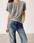 Dressed casually, the person wears "The Crew," a boxy fit gray t-shirt by Hey Gang, made from 100% cotton and enzyme washed for extra softness. They pair it with blue jeans and lean against a plain, light-colored background.