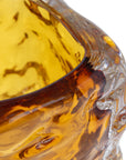 Close-up of the amber 49-704 bryson Vase by The Import Collection, featuring a textured wavy surface. This handcrafted glass vase is translucent, allowing light to emphasize its intricate details against a white background.