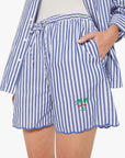 A person wears Mother’s The Drawn FreeBaller Short—lightweight cotton with blue and white stripes, drawstring waist, scalloped hem, pockets, and a small embroidered palm tree on one leg, paired with a matching shirt.