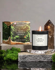 A lit Faire 8 OZ Candle With Lid labeled "Baies Currants Quince" rests on a stone platform next to its box, surrounded by green moss and two tall brown crystals, set against a gray backdrop—a true luxury scented candle experience.