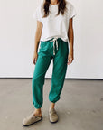 A person stands against a white background in a Hey Gang T-shirt and The Sweatpants from Hey Gang, featuring an elastic waistband, paired with beige slip-on shoes. Their hair is loose, and they face forward.