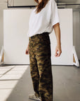 Indoors, a person wears Hey Gang's The Officer Pant in cotton ripstop camouflage, paired with a loose white t-shirt and beige shoes against a minimal white background. The person's face is not shown.