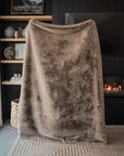 A large, soft brown Cozy Earth Cuddle Blanket with an ultra-plush pile is held up in front of a modern fireplace and dark wall, with decor shelves and a painting visible to the left.