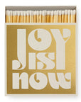 A box of Faire Matches/Square features a gold cover with bold white "JOY IS NOW" text. White match heads are partially visible at the top, reflecting the product’s unique matchbox design.