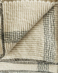The Faire Cozy Stripe Throw is a folded, off-white wool-blend blanket featuring black stitched geometric lines. A turned-up corner reveals the stitching detail on both sides.