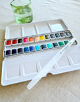 The Faire Watercolor Paint Set w/ Brush, plus an empty mixing palette and a glass of water, are placed on a beige tablecloth.