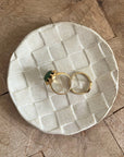 Two gold rings, one with a green stone, rest on Faire’s Ghost Checker Trinket Dish—a round, off-white glazed stoneware dish with a subtle checker pattern—displayed on a wooden surface.
