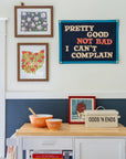 A small kitchen corner with orange mixing bowls, a cookbook, and a tin on a wooden cart. Above, framed wall art and an Oxford Pennant banner read “Pretty Good Not Bad I Can’t Complain.”.