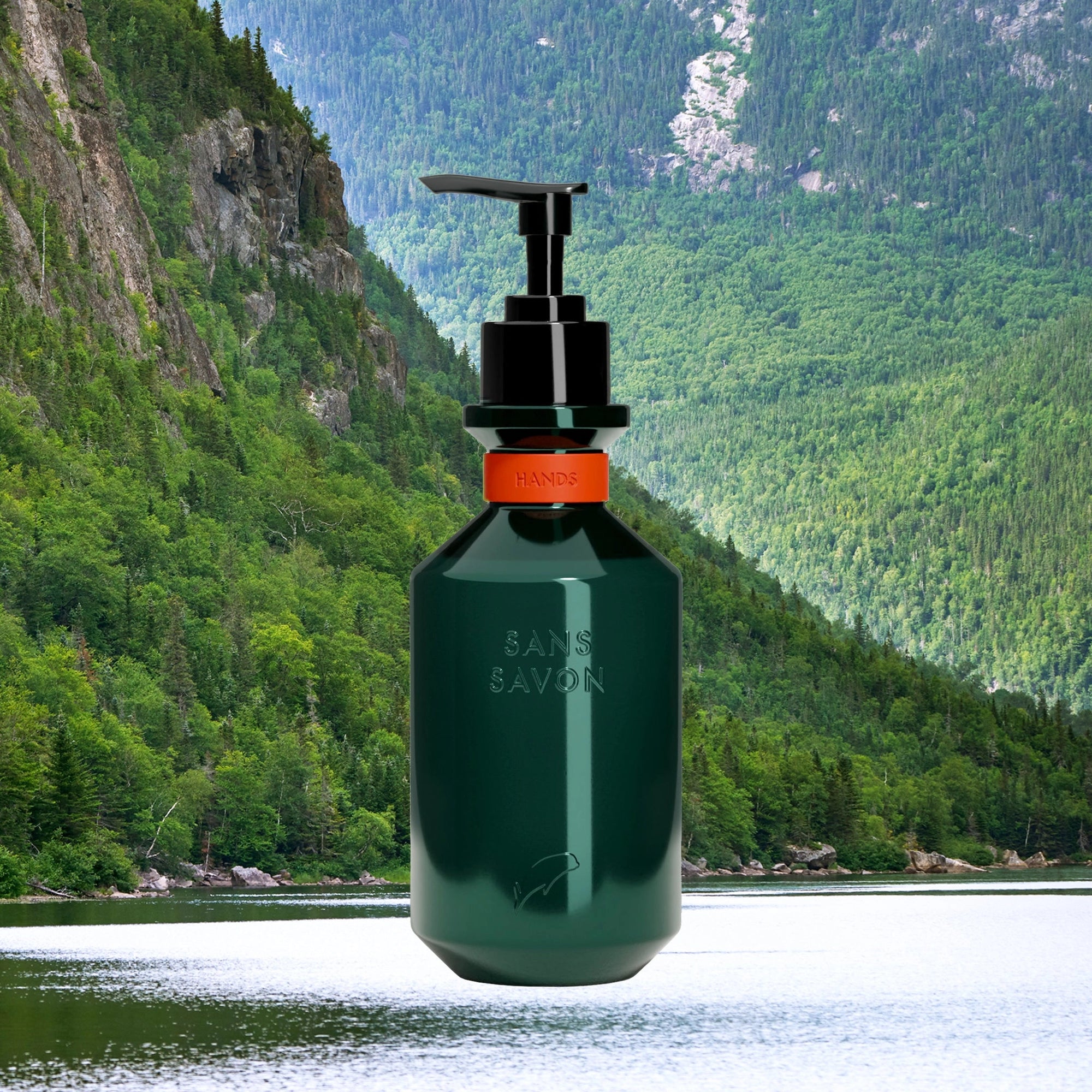 A dark green Faire Hand Wash Charlevoix Boreal dispenser sits before a lush forest, rocky cliffs, and a calm river—emphasizing its microbiome-friendly formula.