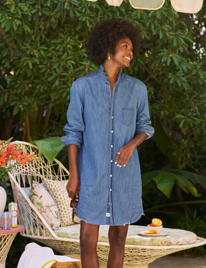 A woman smiles outdoors in the Frank & Eileen Mary Classic Shirtdress - Ruffle, holding sunglasses amid tropical greenery and a patio set with floral cushions and refreshments in the background.