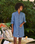A woman smiles outdoors in the Frank & Eileen Mary Classic Shirtdress - Ruffle, holding sunglasses amid tropical greenery and a patio set with floral cushions and refreshments in the background.