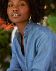 A woman with curly hair and large hoop earrings poses outdoors among green and red foliage, wearing the Frank & Eileen Mary Classic Shirtdress- Ruffle.