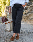 A person stands on a gravel path wearing ankle-length black Frank & Eileen Westport Italian wide leg pants, a white sweater, and brown loafers. They hold a beige tote with leafy branches; behind them is a stone wall and greenery.