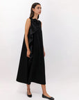 A woman wears the Psophia 2784 Womens Dress, a black mercerized cotton sleeveless midi with a sculptural rosette detail, paired with black flats, standing against a plain white background.