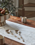 A wooden table with a white runner, two striped Botera Spoons, a bowl of fruit, and ceramic mugs. Blurred stairs and greenery appear in the background.