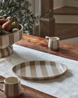 A rustic wooden table features minimalist dinnerware: the beige and white striped Botera Platter by Accent Decor, two matching mugs, and a large fruit bowl on a white runner, with a staircase and leafy plant in the background.