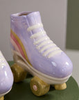 The Accent Decor Rollerskate Planter is a quirky ceramic plant pot painted in pastel lavender, pink, and yellow stripes with beige-yellow wheels—an ideal gift for plant lovers. Displayed on a green surface.