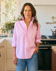 A woman stands in a bright kitchen wearing the Frank & Eileen BARRY Tailored pink and white striped slim fit shirt with rolled sleeves and blue jeans, smiling softly with one hand in her pocket. Plants and a stove are visible in the background.