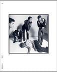 Five men are in a studio; four gaze at a fifth on the floor. This black-and-white image with a wide white border captures Basquiat's raw energy, as featured in "Seeing Loud, Basquiat and Music" by Simon & Schuster Inc.