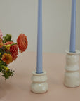 A bouquet of orange and pink flowers sits beside two light blue taper candles in Accent Decor’s Lyam Candleholders, featuring a white reactive glaze, on a pale pink surface for charming handmade decor against a neutral background.