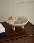 The Accent Decor Walden Bowl, a shallow, speckled ceramic piece with three short legs, sits on a dark wooden surface near books and a light-colored wall—ideal as sculptural home decor.