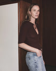 A woman with long hair stands indoors against a wooden wall, wearing the Tabitha Cardigan by Citizens Of Humanity/AGOLDE, paired with light blue jeans and gold jewelry, looking toward the camera with a neutral expression.