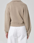 A person is shown from the back, wearing a light brown Citizens Of Humanity/AGOLDE trapeze pullover with a collar and high-waisted white pants against a plain, light gray background.