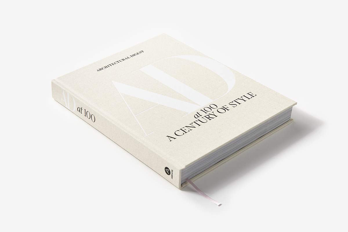 Architectural Digest at 100" by Hachette Book Group is a hardcover book celebrating Architectural Digest’s design legacy, featuring a light beige cover with embossed white "AD" letters and a ribbon bookmark, shown on a white background.