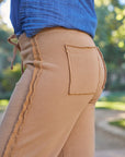 Close-up of a person wearing Frank & Eileen's Bella Sweatpant in tan, featuring 100% cotton fabric, raw edge seams, and a back patch pocket, paired with a tucked-in blue shirt; blurred green outdoor background.